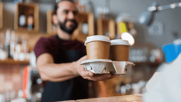 A smiling barista in a German bakery or café handing over two takeaway coffee cups, representing a service sector employee impacted by the Minimum wage up.