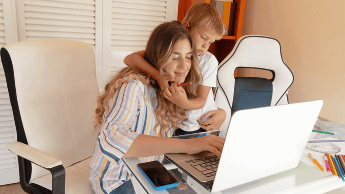 A mother smiles while working on her laptop at home, with her child playing in the background, illustrating the improved work-life balance possible with hybrid work.
