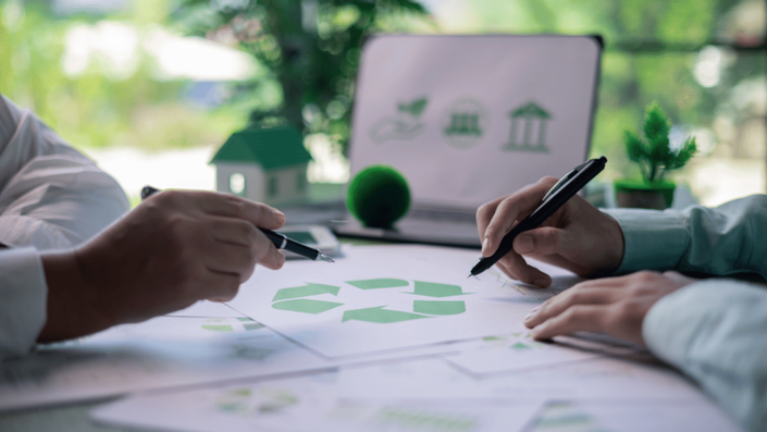 Two professionals discussing a sustainability project at a desk, reviewing documents with a green recycling symbol. The workspace includes eco-friendly decorations and a laptop displaying environmental icons. A representation of sustainable ventures in business strategy and development.