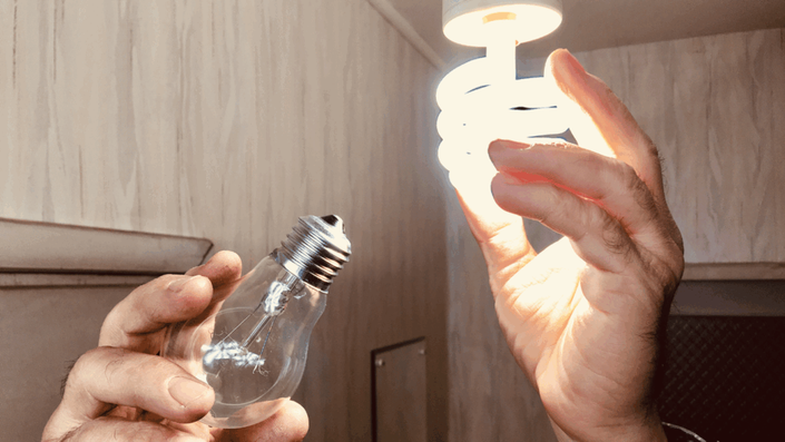 A hand holding an old incandescent light bulb next to another hand holding a new, glowing energy-efficient spiral light bulb, symbolizing a switch to more sustainable options for Everyday Savings.