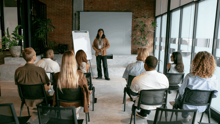 A speaker stands in front of a whiteboard and screen, addressing a diverse audience seated in rows, illustrating a presentation or workshop setting for incubators and accelerators that support business ideas for young founders.
