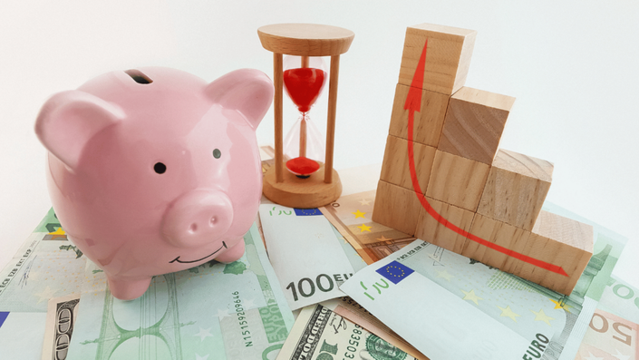 A piggy bank, hourglass, and growing investment blocks on top of euro and dollar bills, representing diversification in your investment portfolio and financial growth.