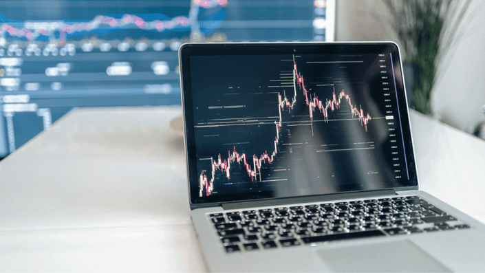 A laptop screen showing detailed financial candlestick charts, with a larger monitor displaying more data in the background, representing analysis of Cryptocurrency Trends.