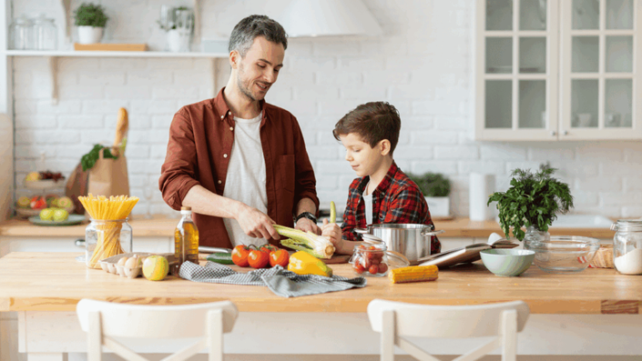 A father and son happily cooking together in a bright kitchen, preparing fresh vegetables, illustrating the benefits of meal planning and home cooking for Everyday Savings and a healthy lifestyle.