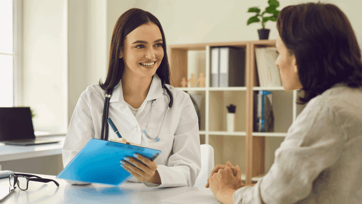 A smiling female doctor holding a clipboard and discussing Health Insurance options with a patient in a modern office setting.